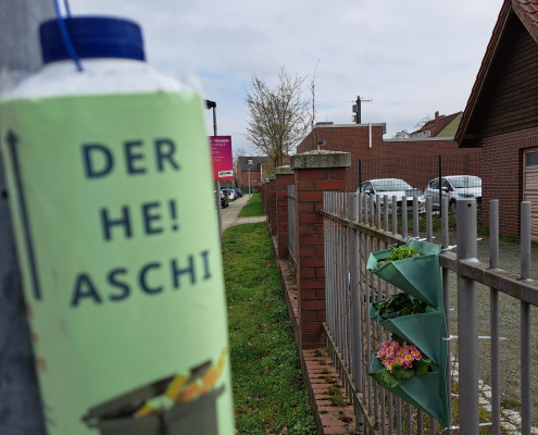 Eine grün angemalte leere Milchtütte mit dem Schriftzug "der HE! Ascher" an einen Zaun gehangen